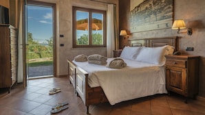 A bedroom with a bed, a nightstand, a lamp, a painting, and a view through a window. - Casanova - Panoramic Rooms and Suites (San Quirico d'Orcia)