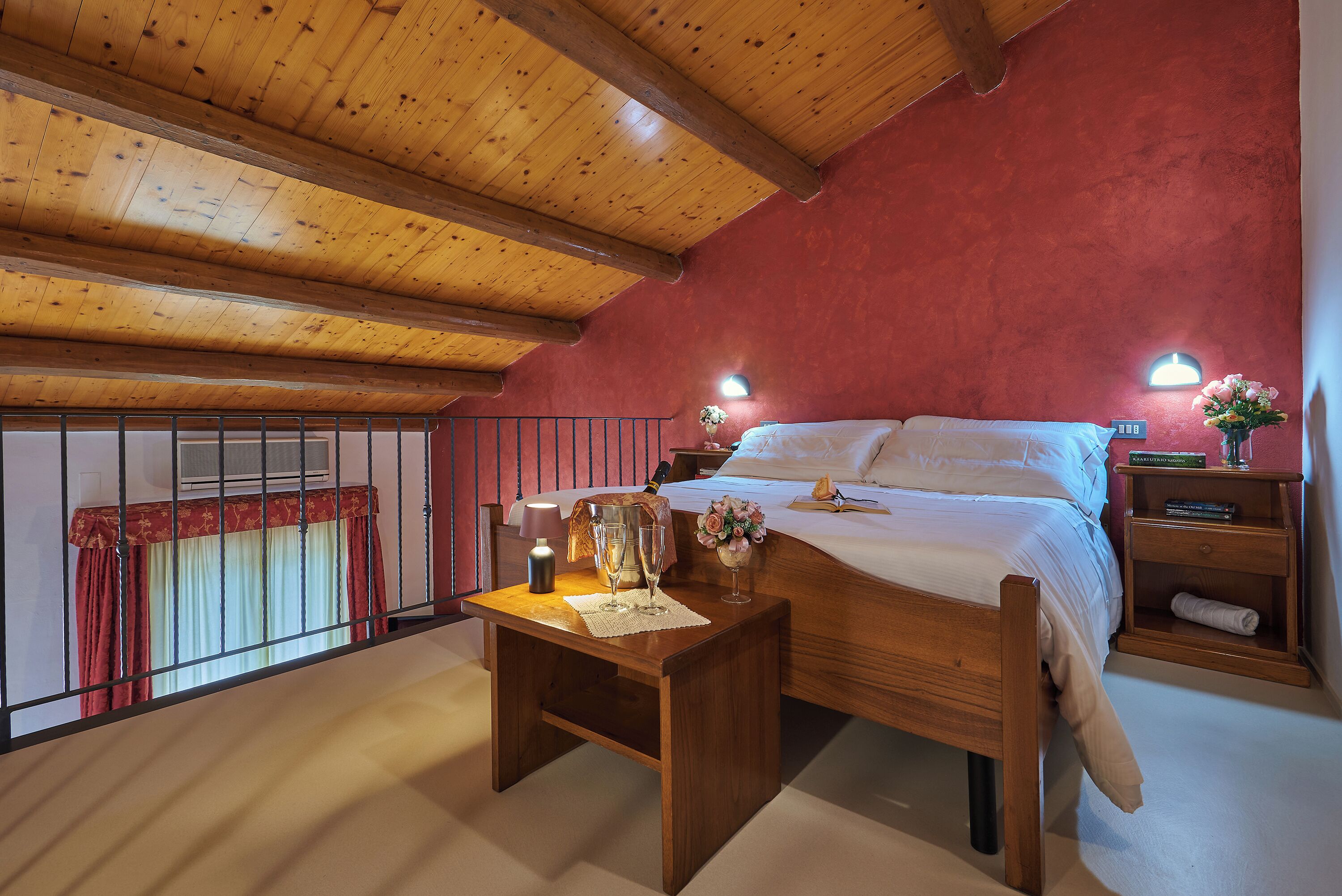 Un dormitorio con techo de madera, cama con sábanas blancas, mesita de noche con lámpara y ventana con cortinas rojas.