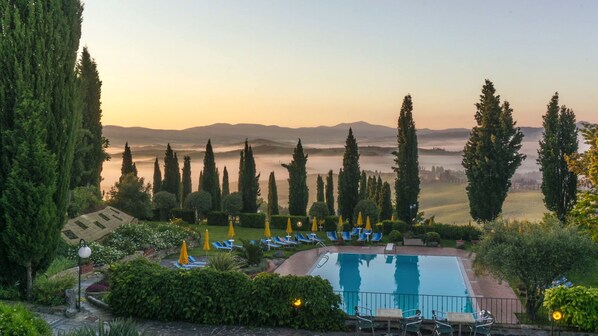 A pool area with lounge chairs and umbrellas, surrounded by tall cypress trees and a view of distant hills. - Casanova - Panoramic Rooms and Suites (San Quirico d'Orcia)
