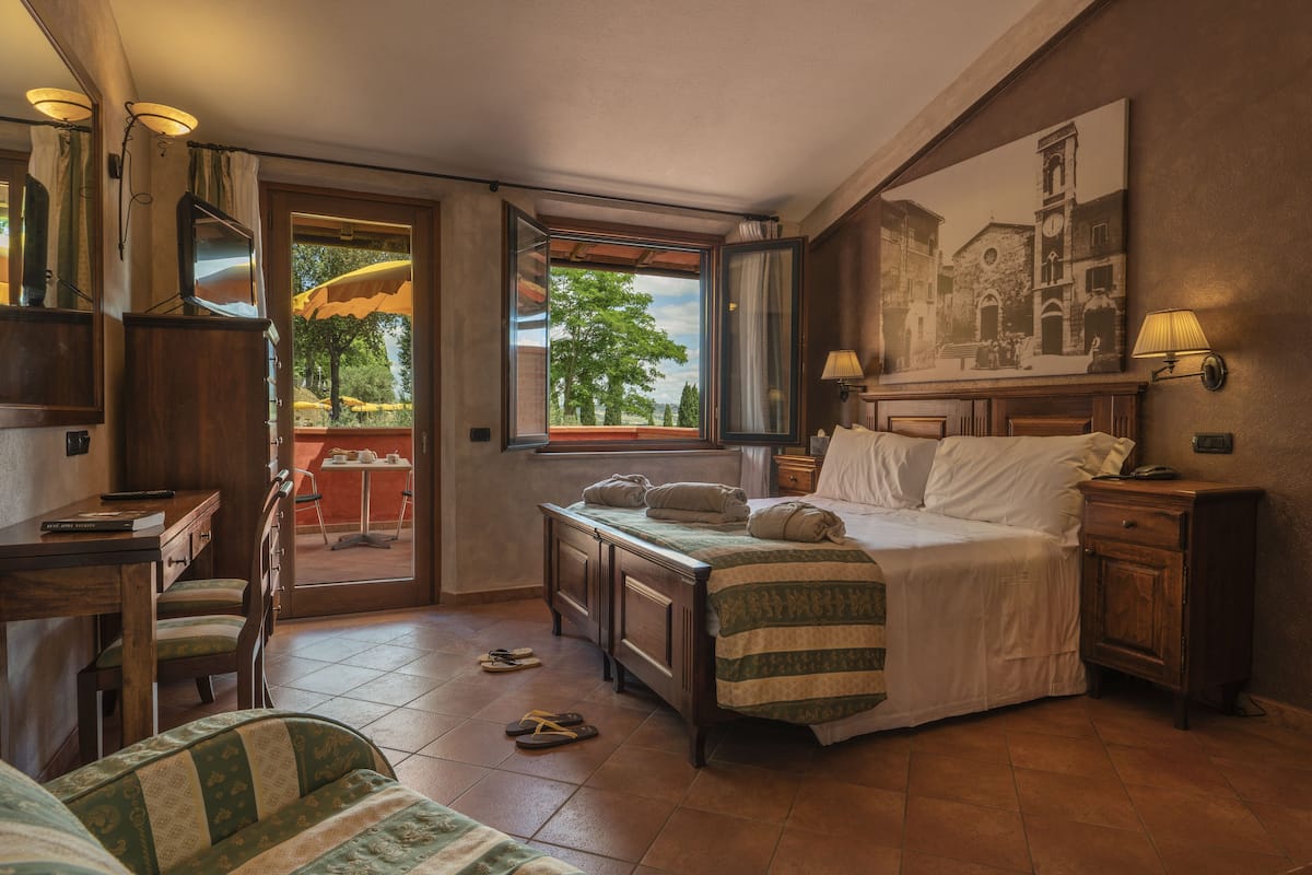 A bedroom with a bed, a desk, a chair, a window with a view of trees, and a painting on the wall.