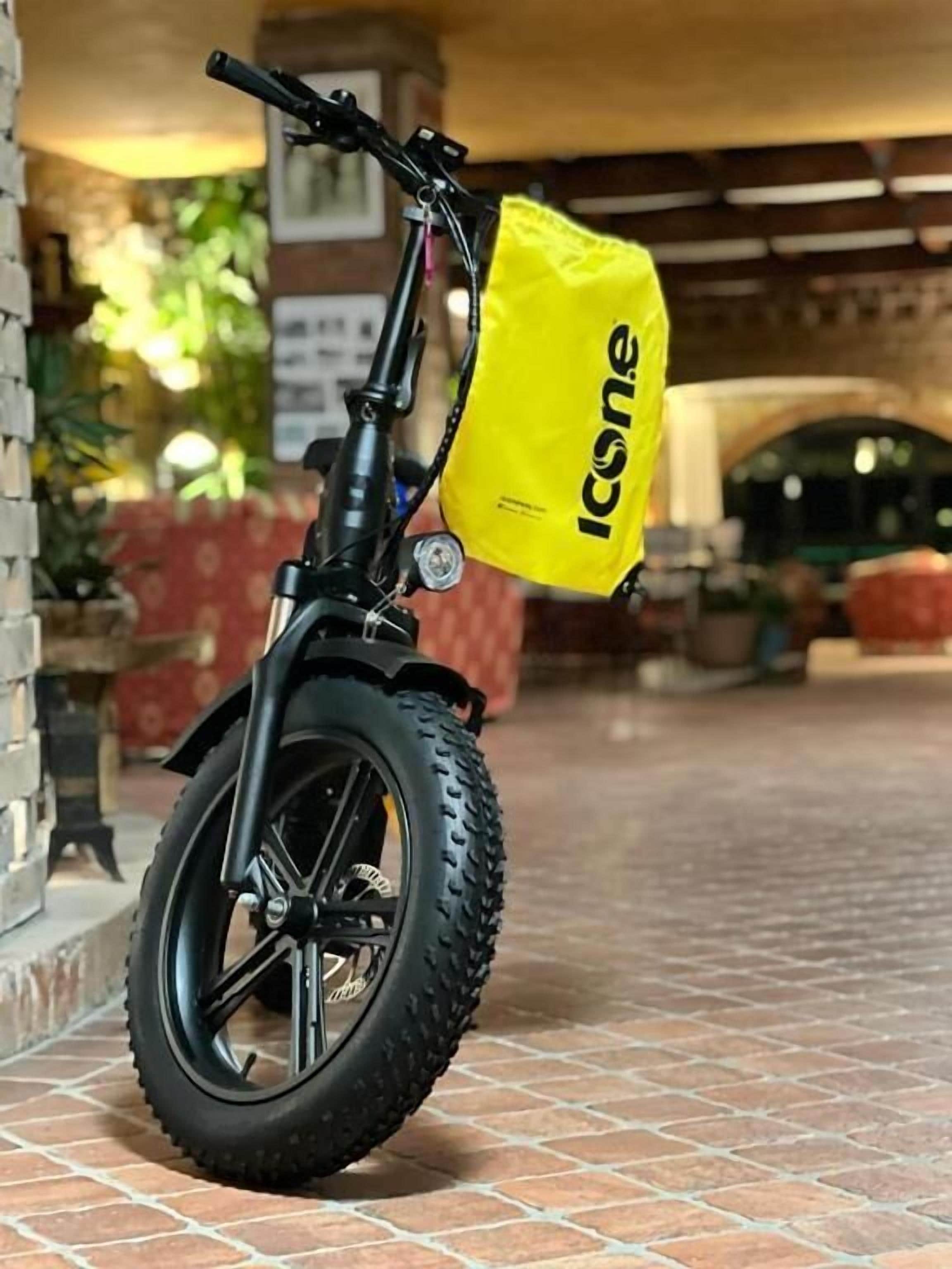 A black electric scooter with a yellow bag labeled "Icone" attached to it, parked on a tiled pavement.