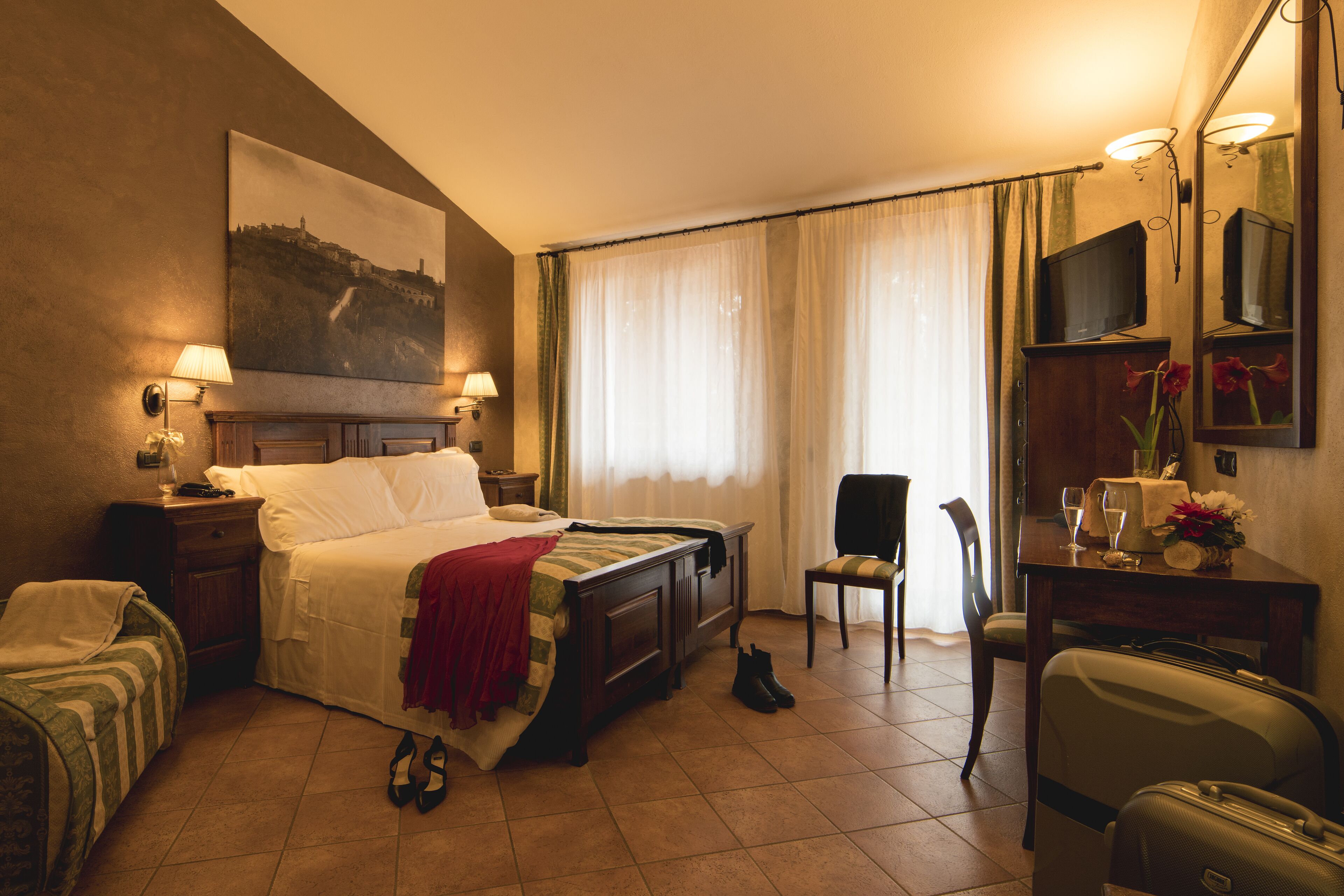 Habitación de hotel con cama, una silla, un escritorio con espejo, televisor y maleta.