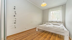 A bedroom with a wooden floor, a bed with white bedding, a window with sheer curtains, and a hanging light fixture. - Victus Apartamenty - Poggio (Sopot)