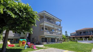 A multi-story building with a balcony, a small play area with toys, and a paved walkway. - By the Sea (Kassandra)