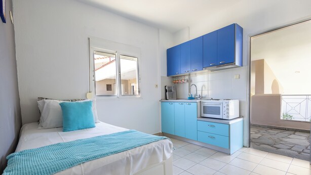 A bedroom with a bed, a window, and a kitchenette with blue cabinets and a microwave. - By the Sea (Kassandra)