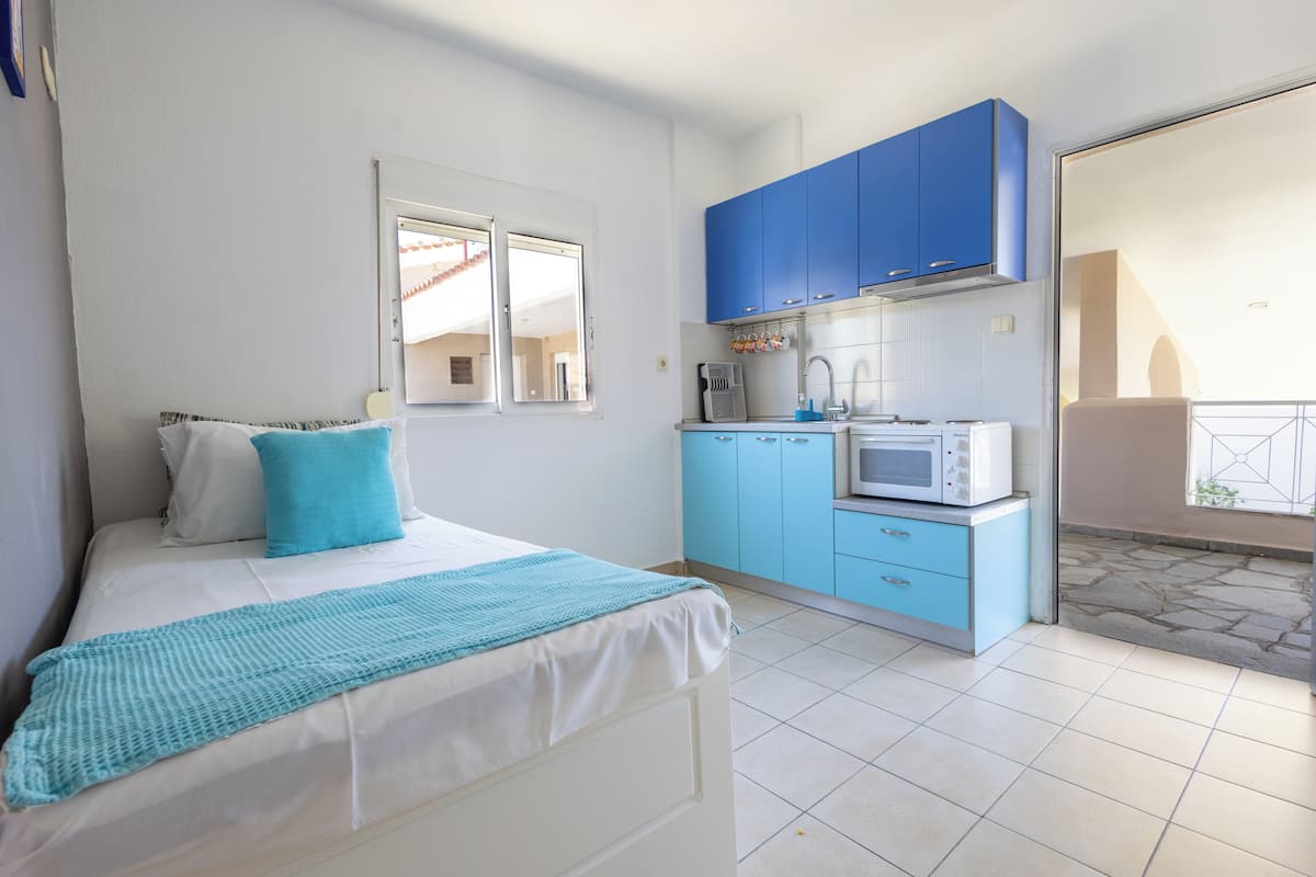 A bedroom with a bed, a window, and a kitchenette with blue cabinets and a microwave.