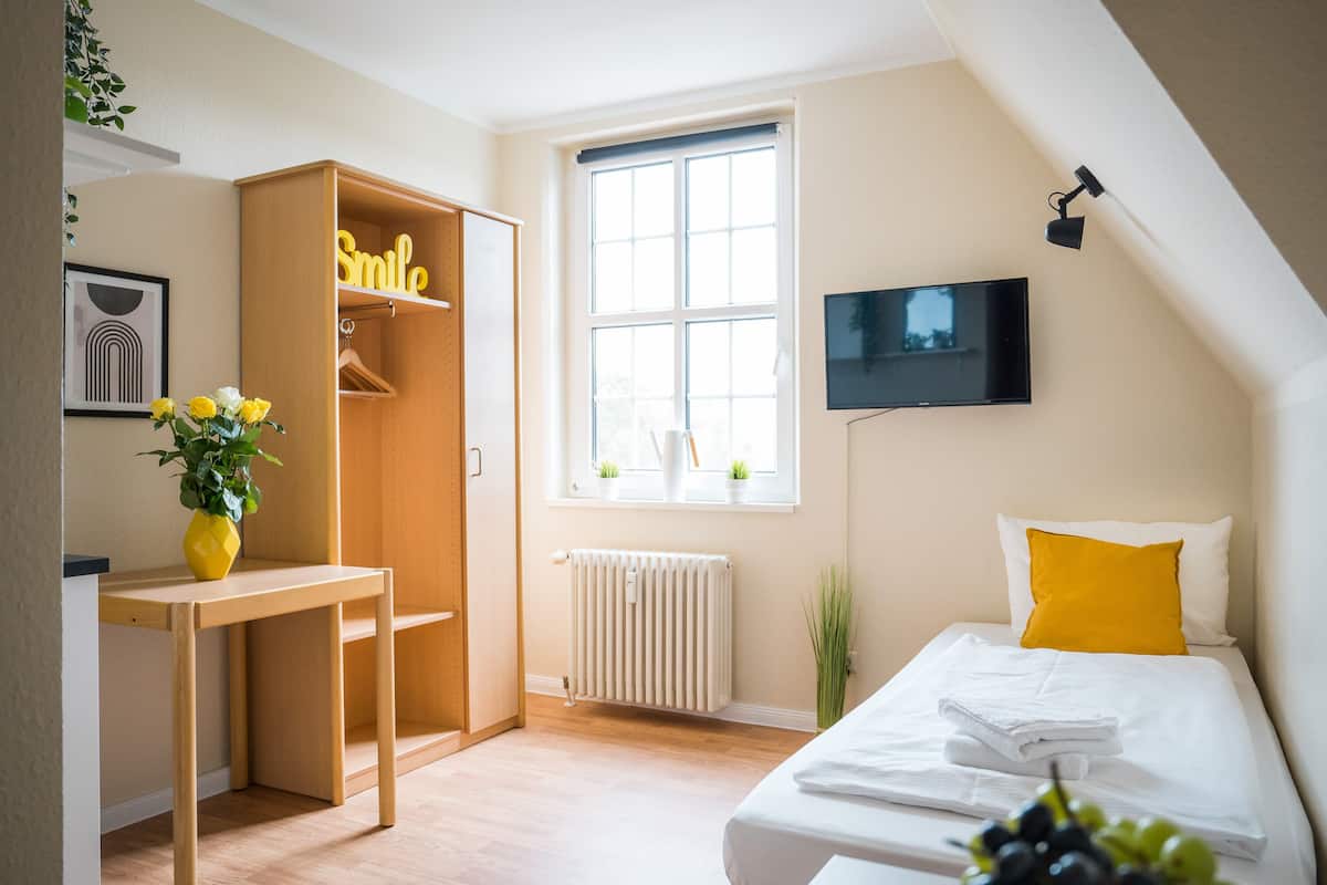 Quarto com cama, escrivaninha, guarda-roupa, radiador, televisão e janela com plantas.