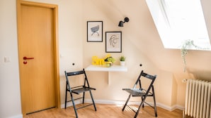 Une pièce avec une porte en bois, deux chaises noires, une étagère blanche sur laquelle se trouve un saladier rempli de fruits et une plante, et des œuvres d’art encadrées accrochées au mur.