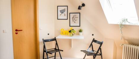 A room with a wooden door, two black chairs, a white shelf with a bowl of fruit and a plant, and framed artwork on the wall.