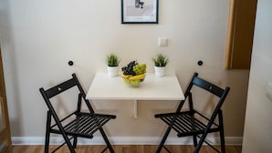 Un coin repas avec une table blanche, deux chaises noires, un tableau encadré au mur et un panier de fruits sur la table.