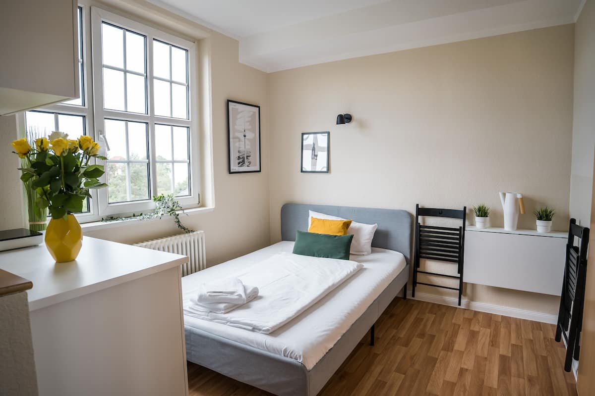 Quarto com cama, escrivaninha, radiador, janela e parede com duas quadros.