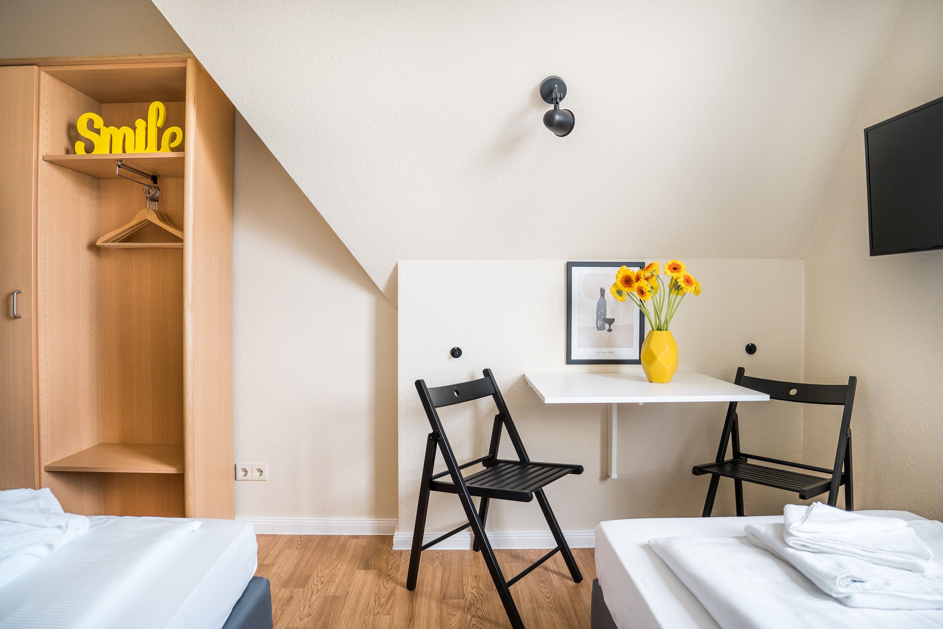 Un dormitorio con una cama, dos sillas negras, un escritorio con un jarrón de flores, una lámpara colgante y un armario con un letrero que dice 'Smile'.