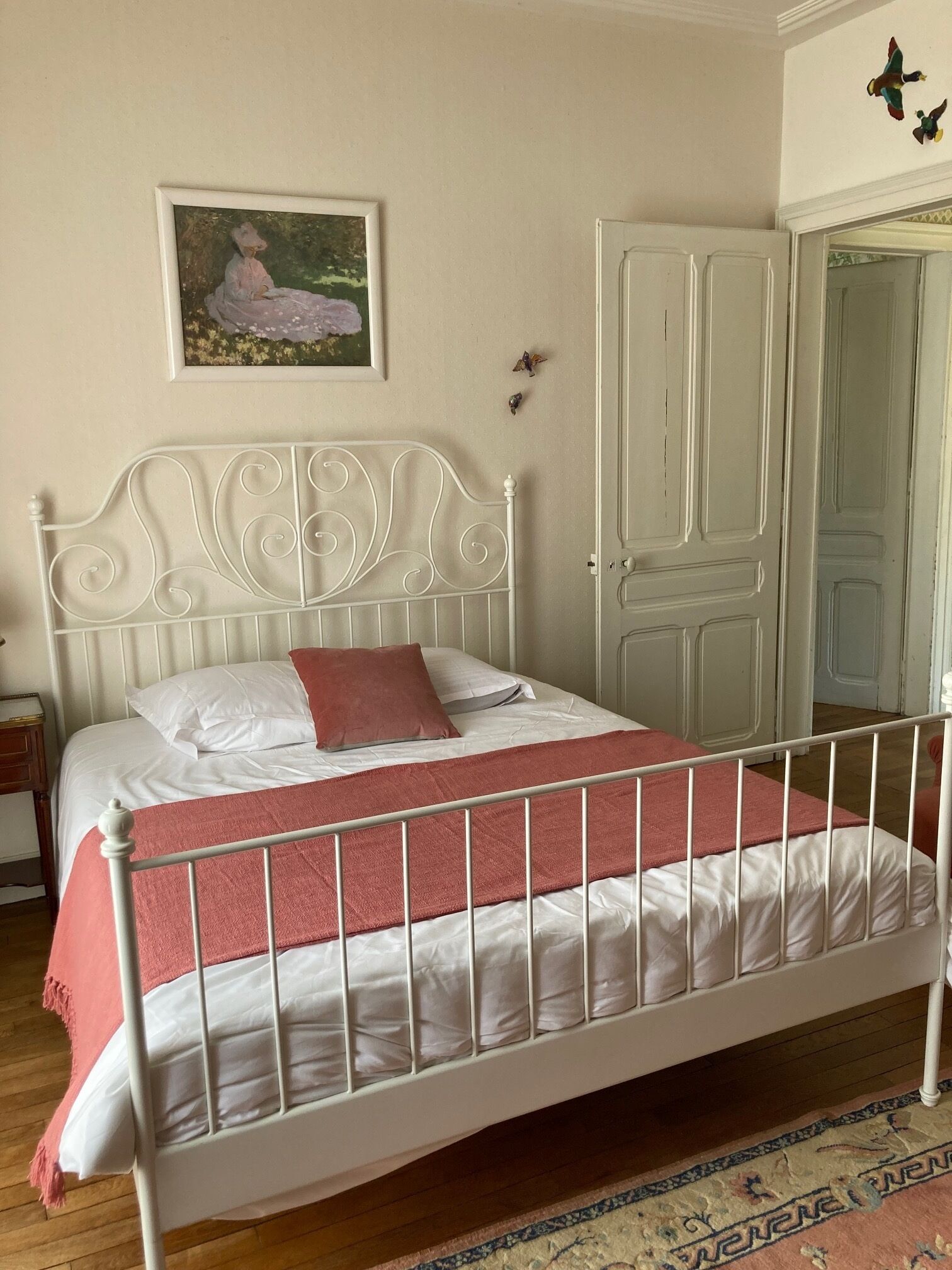 A neatly made bed with a white metal frame and a pink blanket, a white door, and a framed painting on the wall.