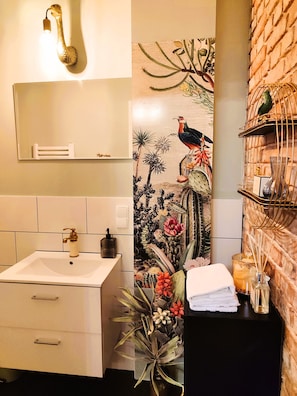 Une salle de bain avec un lavabo blanc, un miroir, un luminaire fixé au mur, un panneau décoratif et une étagère contenant des articles de toilette.