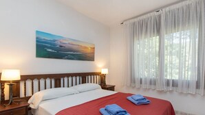 A hotel room with a bed, two bedside lamps, a painting of a sunset over the ocean, and a window with sheer curtains.