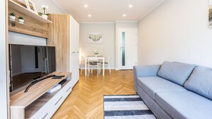 A modern living room with a flat-screen TV, a grey sofa, a wooden herringbone floor, and a dining area with a white table and chairs.
