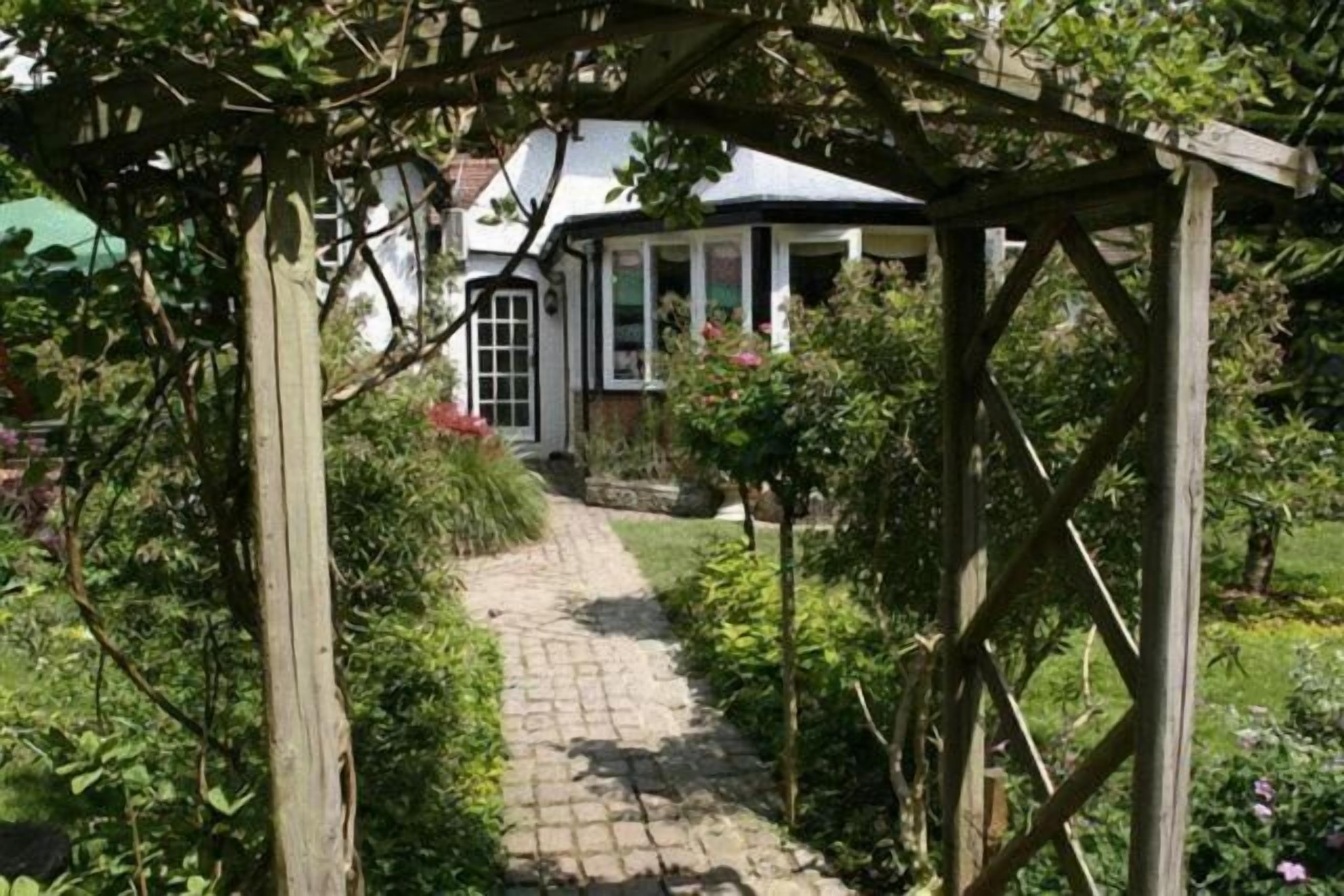 A garden pathway leading to a white building with a black roof, surrounded by lush greenery and a wooden pergola structure.