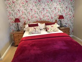 Un dormitorio con papel tapiz floral, una cama de madera, dos mesitas de noche con lámparas y una manta roja.