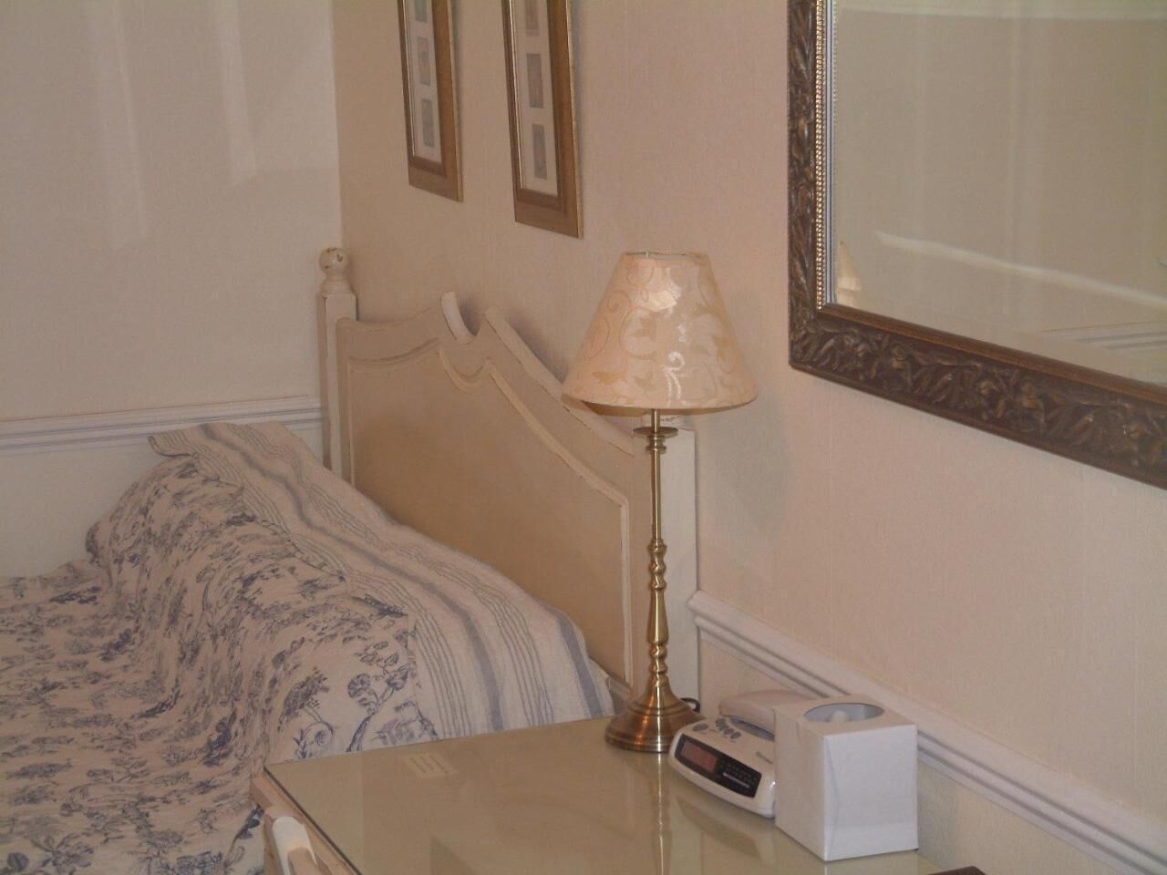 Un lit bien fait, recouvert d’une couverture à motifs floraux, une table de chevet avec une lampe, une horloge numérique et un miroir accroché au mur.