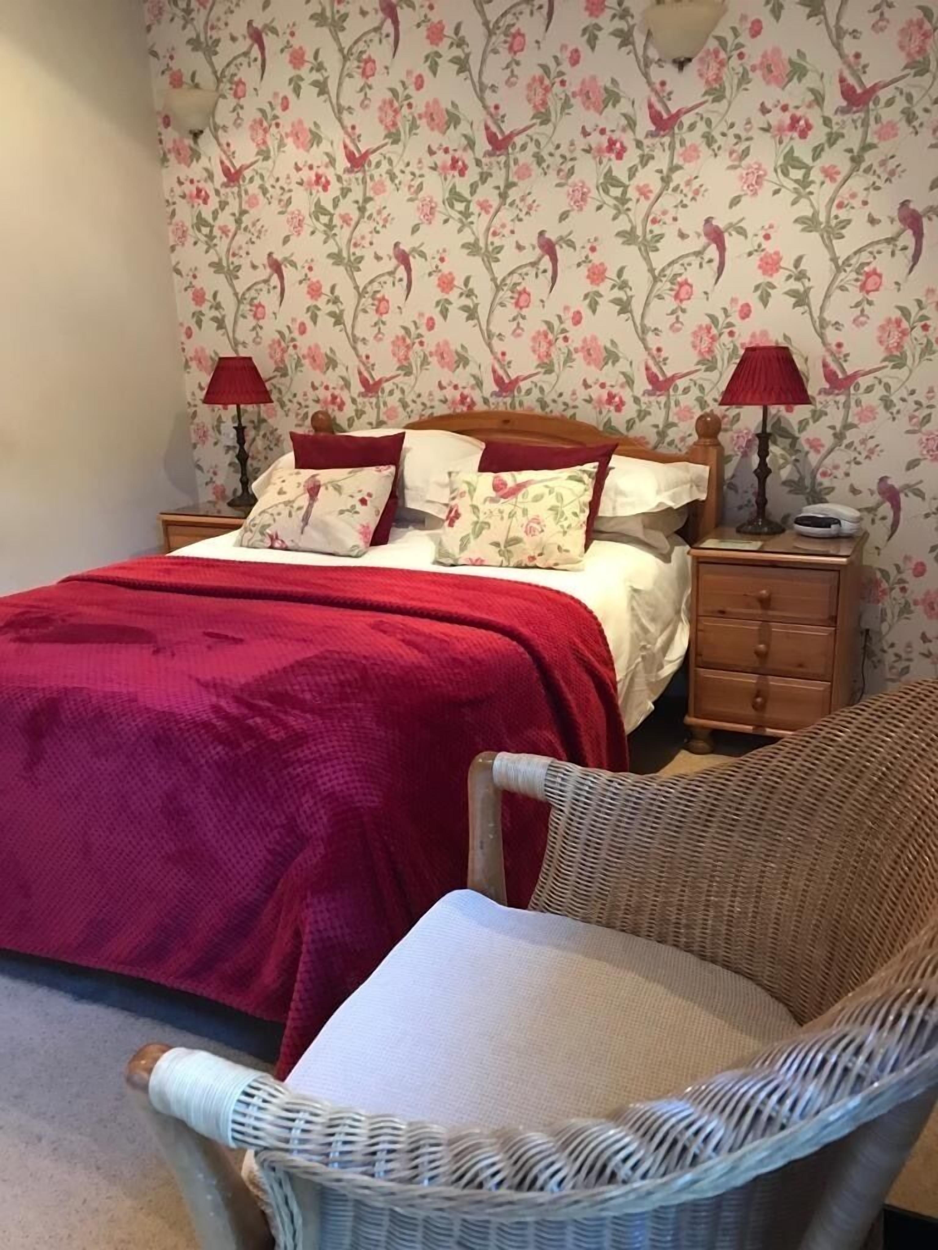Un dormitorio con papel tapiz floral, colcha roja, dos lámparas de noche, mesita de madera y una silla de mimbre.