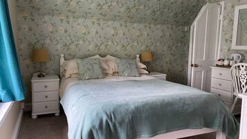 A bedroom with floral wallpaper, a bed with a green blanket, two bedside lamps, a white dresser, and a chair.