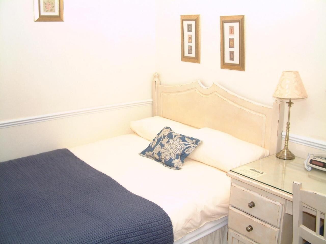 Een opgemaakt bed met een blauwe deken en een bloemenkussen, een nachtkastje met een lamp, en een klein ladeblok.