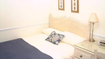 Een opgemaakt bed met een blauwe deken en een bloemenkussen, een nachtkastje met een lamp, en een klein ladeblok.