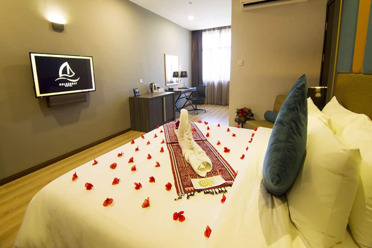 A hotel room with a bed adorned with rose petals, a desk with a chair, a television mounted on the wall, and an air conditioning unit.