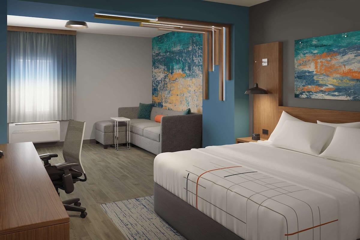 Ein modernes Hotelzimmer mit einem großen Bett, einem Schreibtisch, einem Stuhl und einem Sofa. An den Wänden hängen zwei Gemälde und ein Fenster mit Vorhängen.