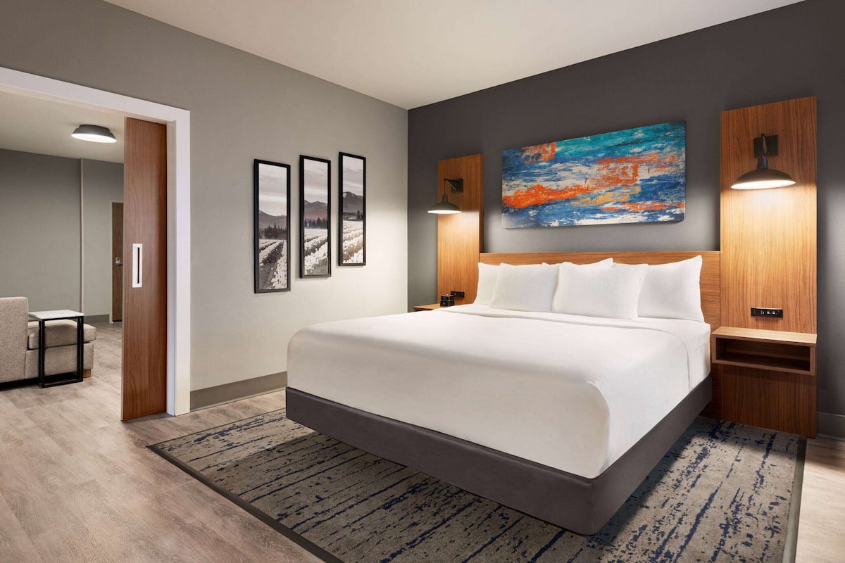 Ein modernes Hotelzimmer mit einem großen Bett, einem Holzkopfende, einem Bild an der Wand und einem kleinen Tisch mit einem Stuhl.