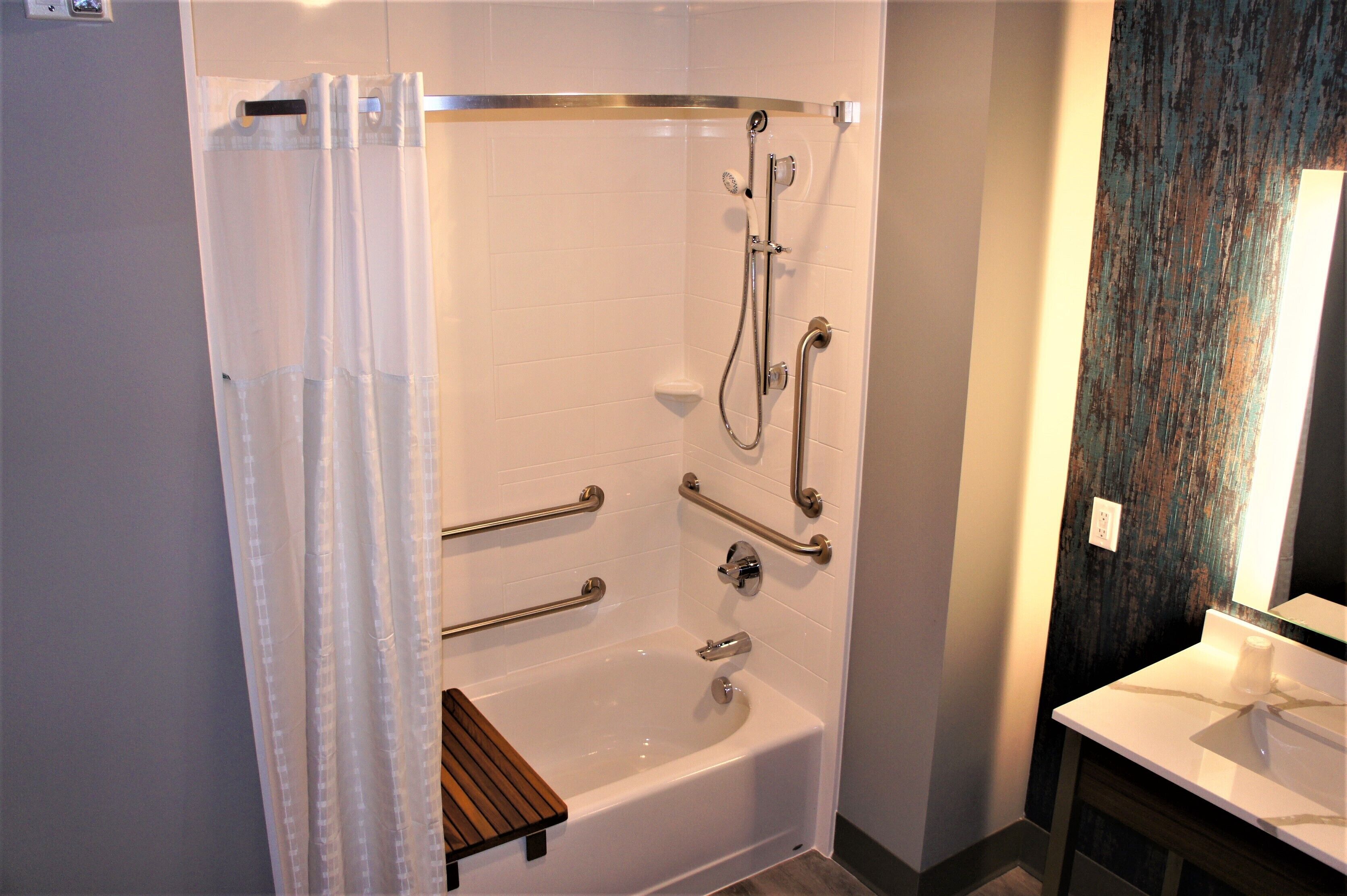 A walk-in shower with grab bars, a wooden shelf, and a white bathtub.