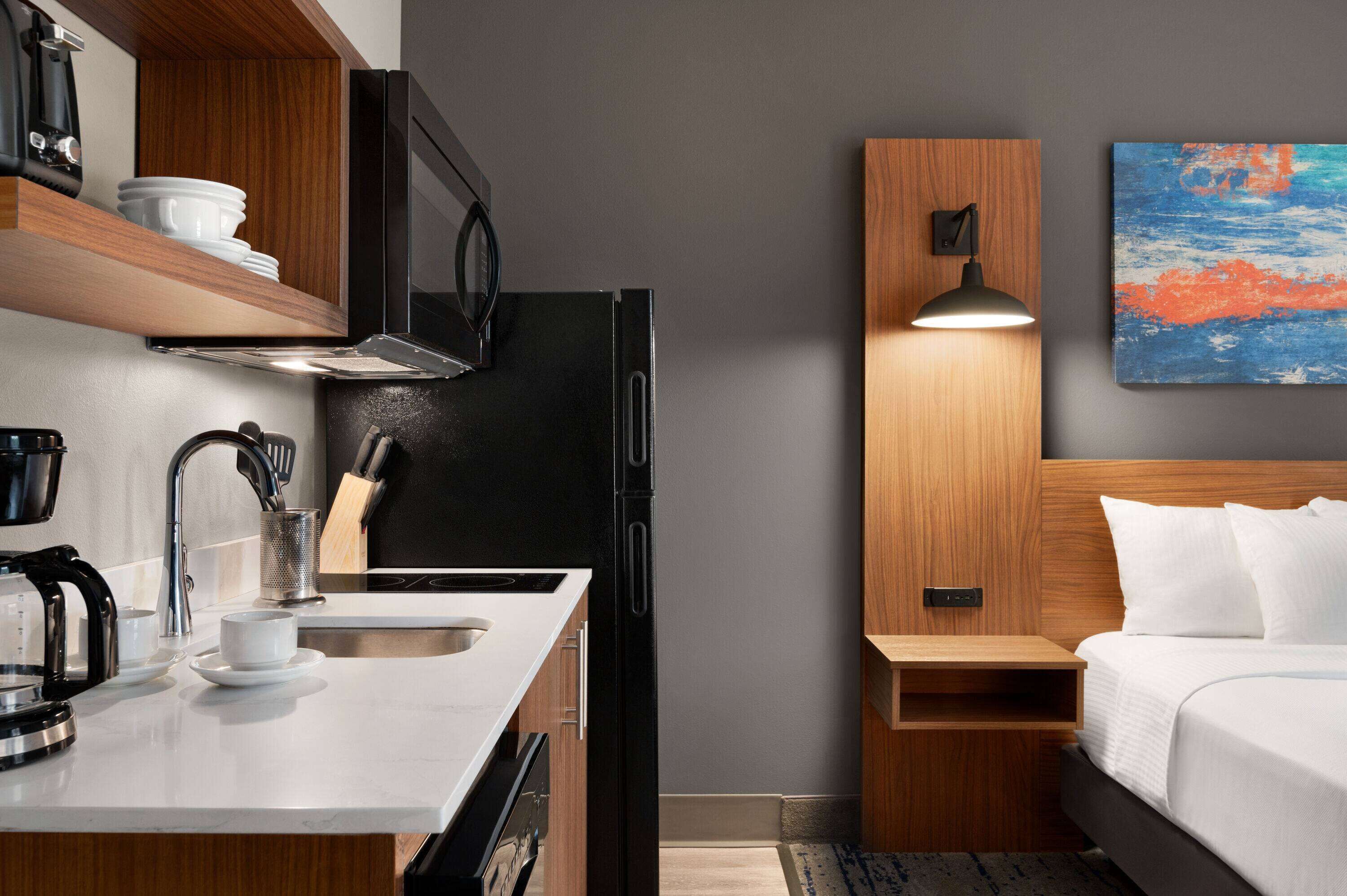 A compact hotel room with a kitchenette, a bed, a bedside table, and a painting on the wall.