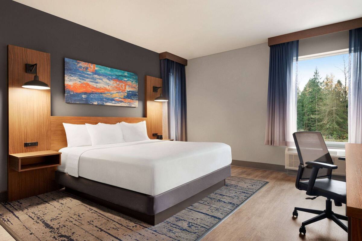 Ein Hotelzimmer mit einem großen Bett, einem Schreibtisch mit Stuhl, einem Bild an der Wand und einem Fenster mit Vorhängen.