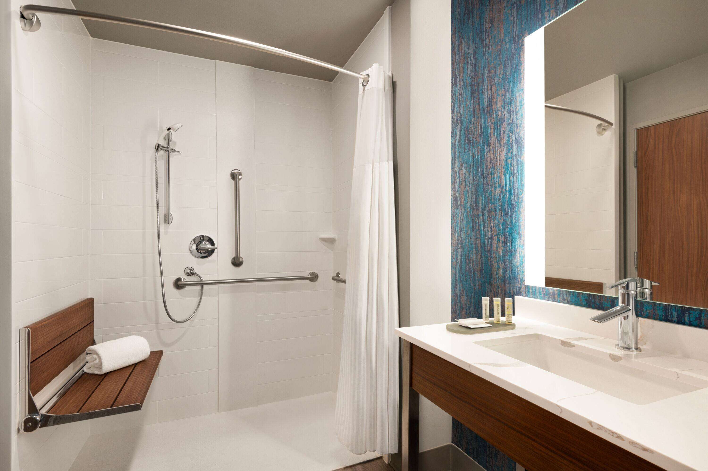 A modern bathroom with a walk-in shower, grab bars, a bench with a towel, a sink with a mirror, and a wooden vanity.