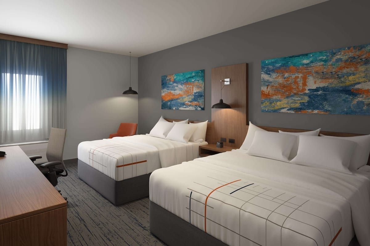 Ein Hotelzimmer mit zwei Betten, einem Schreibtisch und einem Stuhl. An den Wänden befinden sich zwei abstrakte Gemälde.