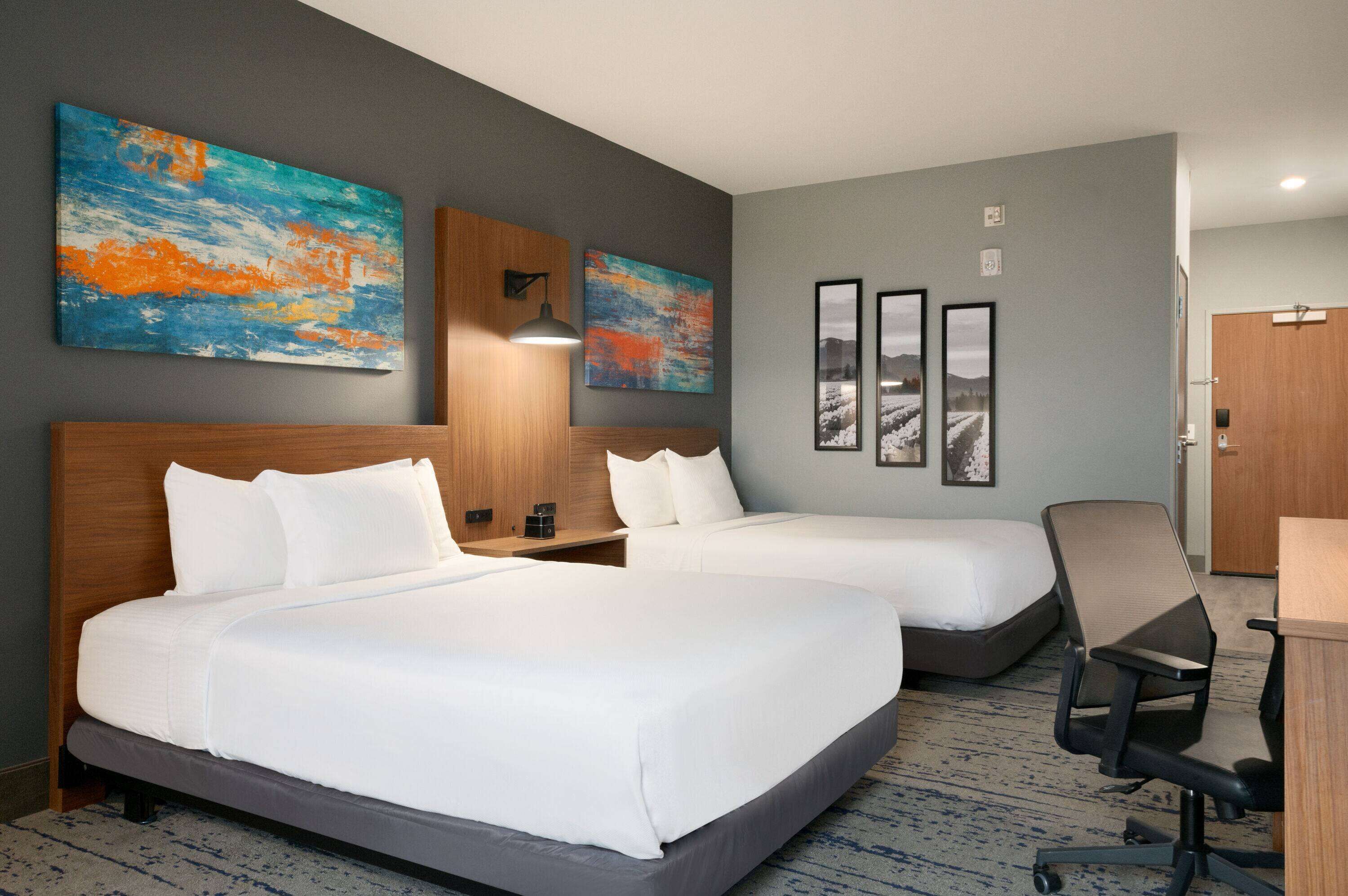 Ein Hotelzimmer mit zwei Betten, einem Schreibtisch und einem Stuhl. An den Wänden hängen zwei Gemälde und drei gerahmte Bilder.