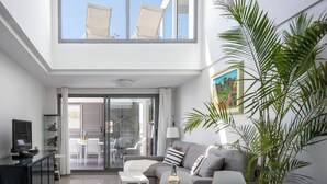 Ein modernes Wohnzimmer mit einer Couch, einem Couchtisch und einem Fernseher auf einem Schrank. Ein großes Fenster und eine Pflanze stehen in der Ecke.
