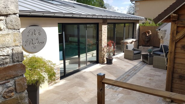 Un bâtiment moderne avec une façade en pierre, une porte en verre et une terrasse en bois aménagée avec du mobilier d’extérieur.