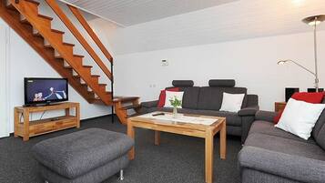 Een woonkamer met een trap, een televisie op een meubel, een bank, een poef en een salontafel.