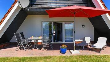 Een terras buiten met een eettafel, stoelen, een rode parasol en een schotelantenne.