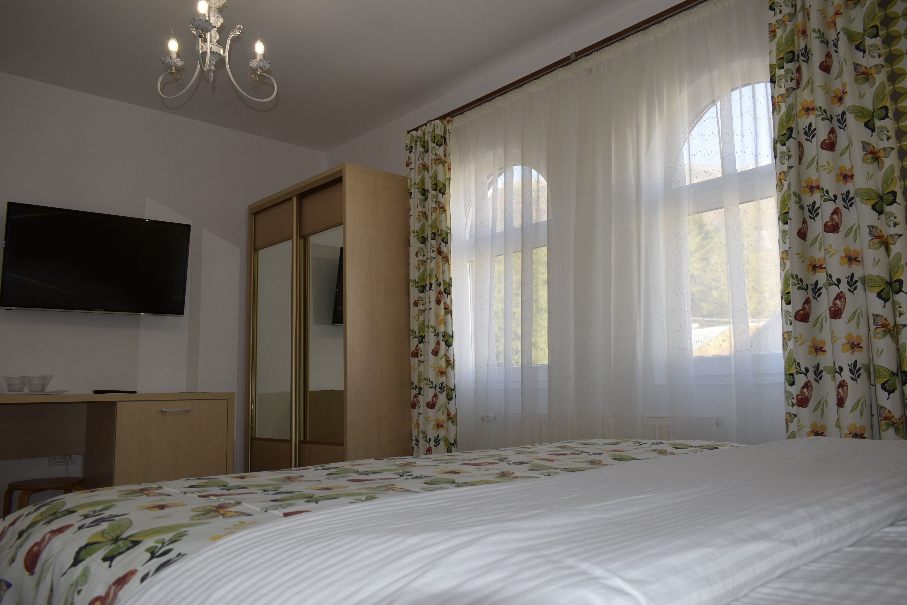 Een slaapkamer met een bed, een aan de muur bevestigde televisie, een kledingkast en een raam met bloemen gordijnen.