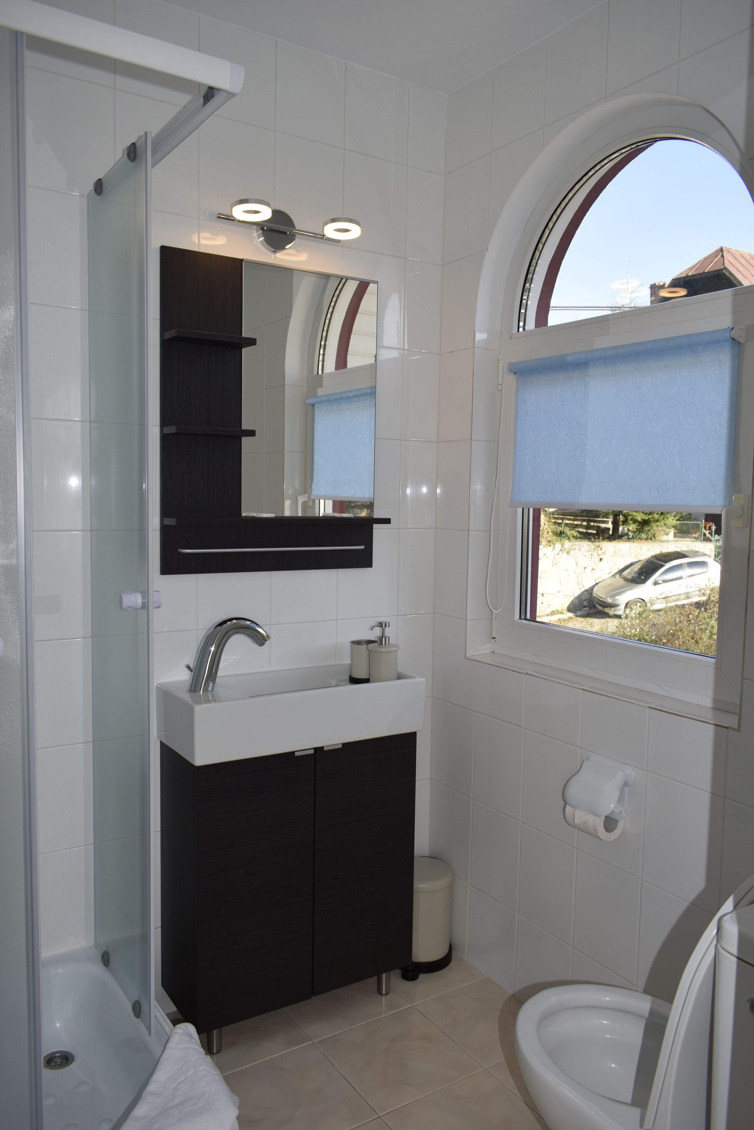 Ein modernes Badezimmer mit einer gläsernen Duschkabine, einem weißen Waschbecken mit dunklem Unterschrank, einem großen Spiegel und einem Fenster mit Blick auf einen geparkten Wagen draußen.