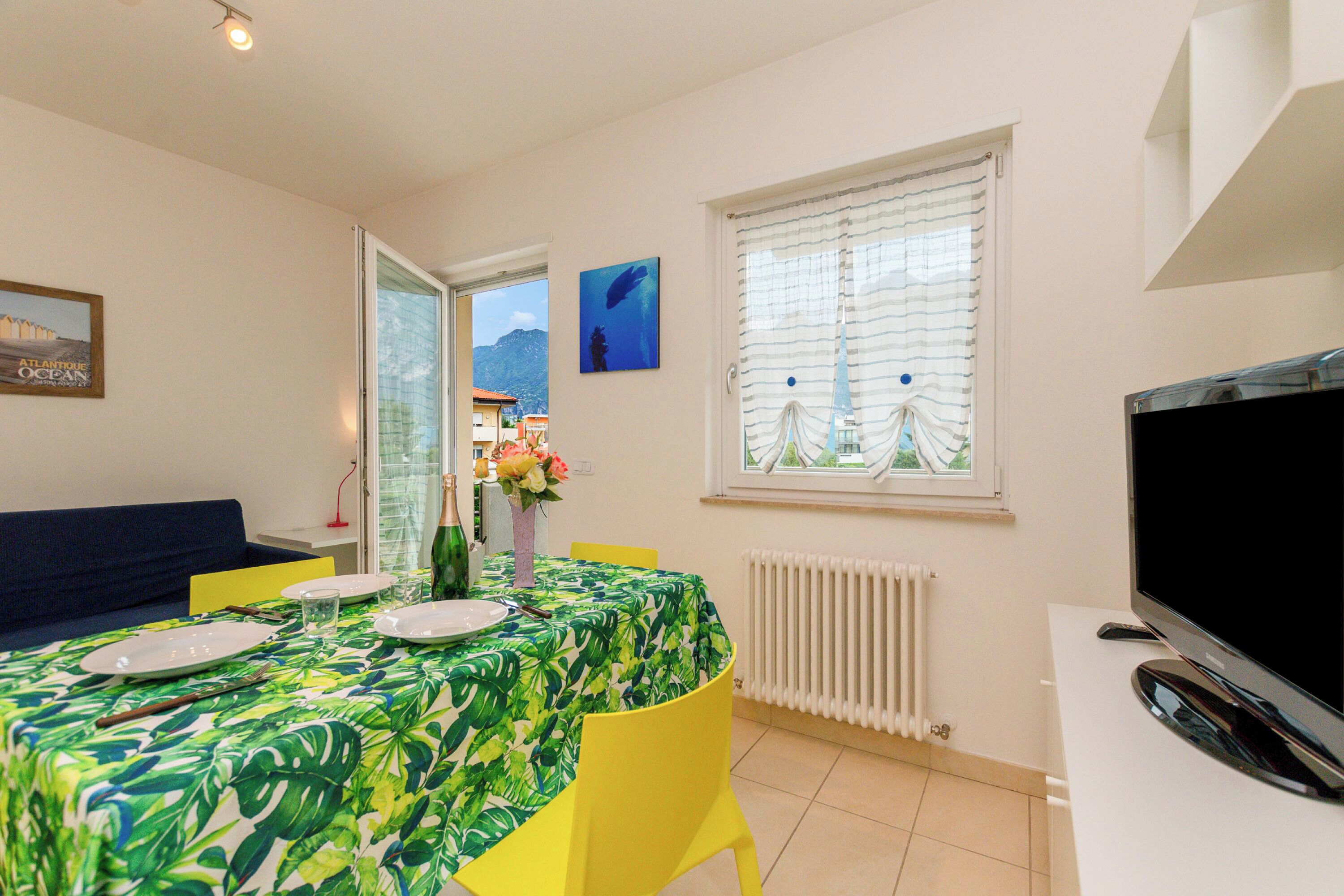 Un área de comedor con una mesa preparada para dos, un sofá, un televisor y una ventana con vistas a edificios y montañas.