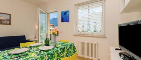 Un área de comedor con una mesa preparada para dos, un sofá, un televisor y una ventana con vistas a edificios y montañas.