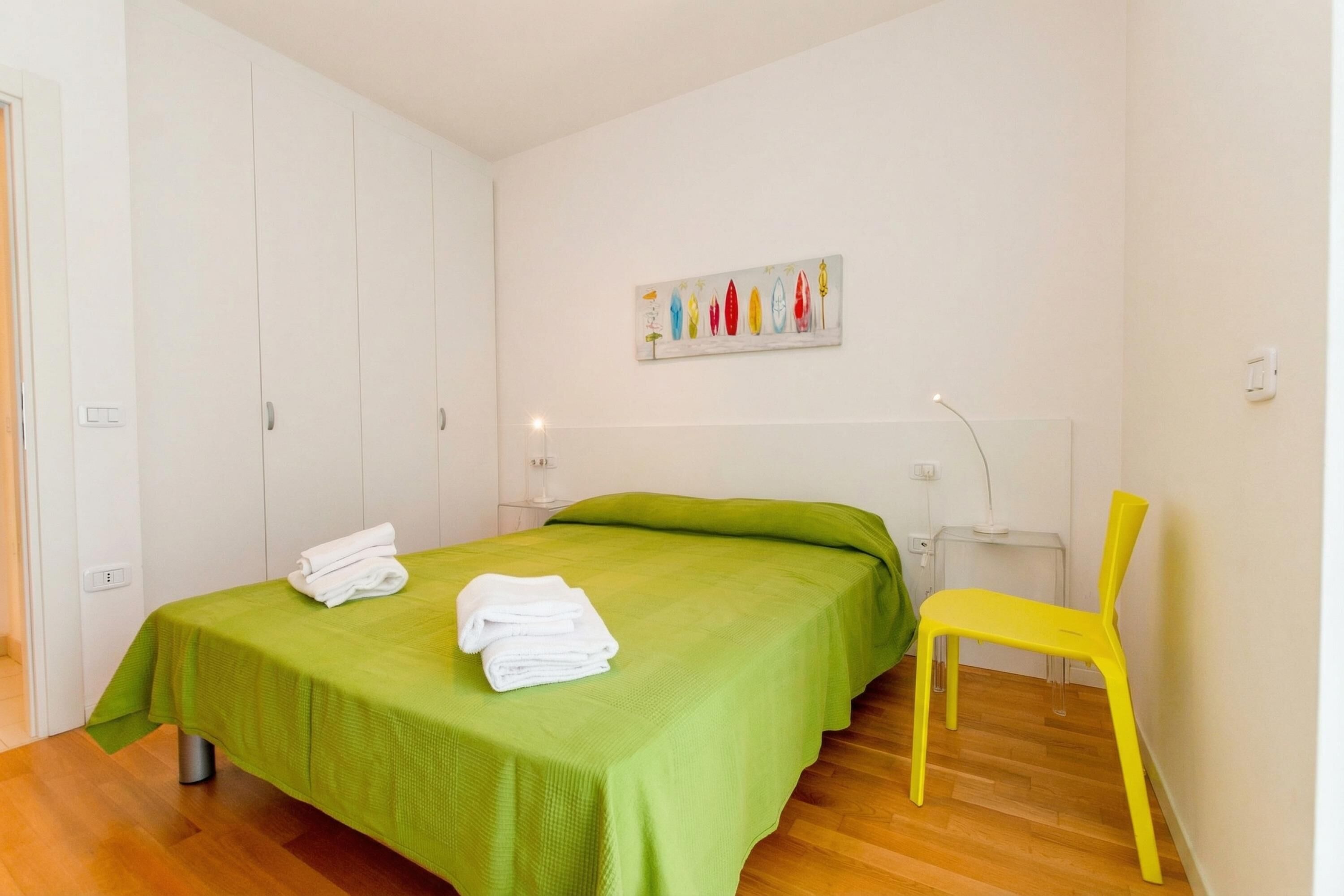 A bedroom with a green bedspread, a white towel, a yellow chair, and a painting on the wall.