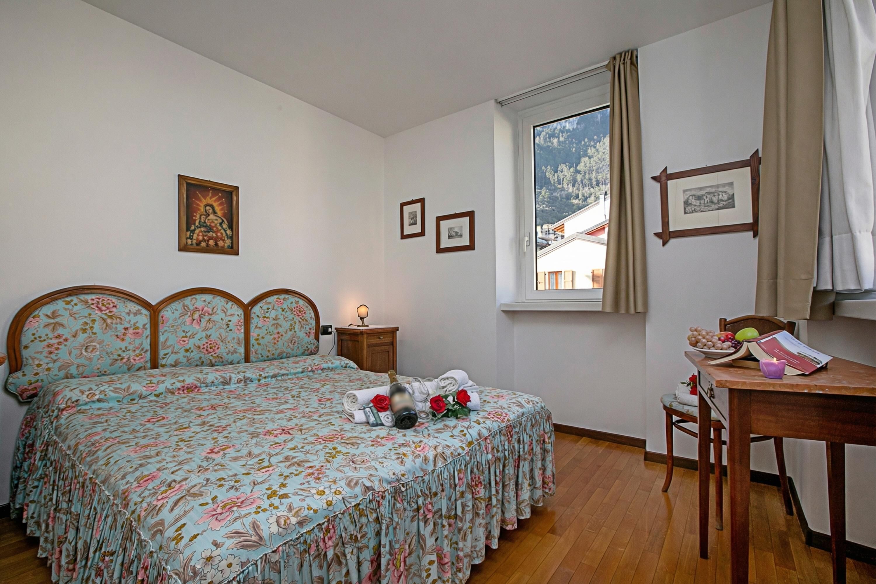 A bedroom with a floral bedspread, a wooden nightstand with a lamp, a desk with a chair, and a window with a view of a building and mountains.
