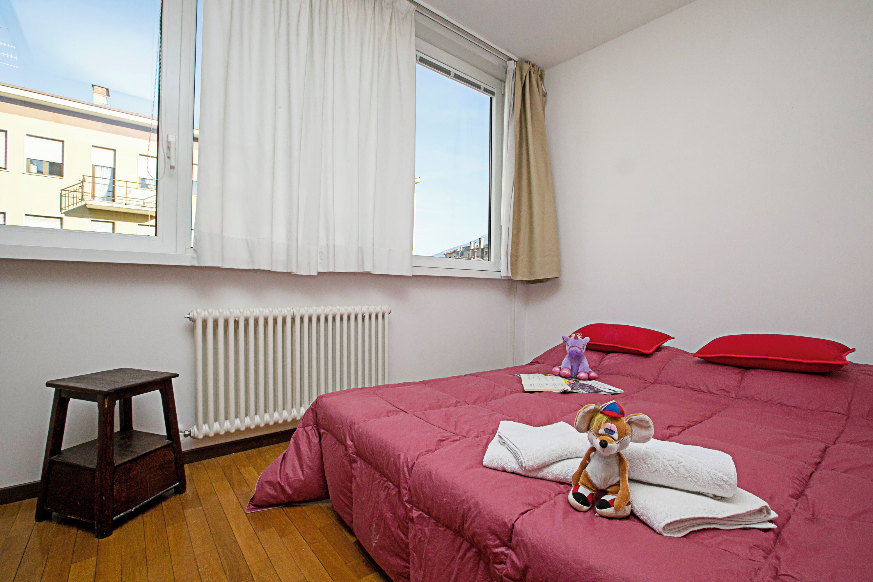 A bedroom with a bed, a window with curtains, a radiator, a wooden stool, and a stuffed toy.