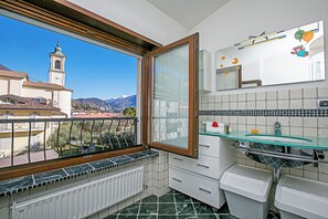 A bathroom with a view of a church and mountains, a window, and a tiled floor. - Paulie Apartment (Riva del Garda)