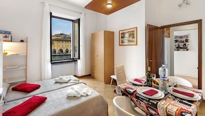 A hotel room with two beds, a dining table set for two, a window with a view of buildings, and a wardrobe.
