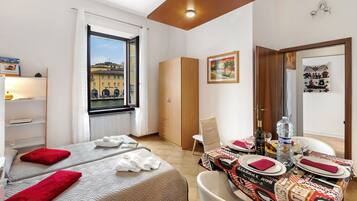 Habitación de hotel con dos camas, una mesa de comedor para dos personas, un ventanal con vistas a edificios y un armario.